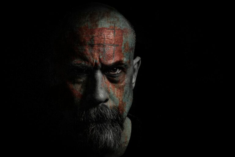 Intense portrait of a bald man with facial tattoos in dark, moody lighting.