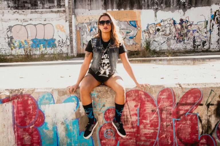 Young woman in stylish attire sitting on graffiti-covered wall in a cityscape.