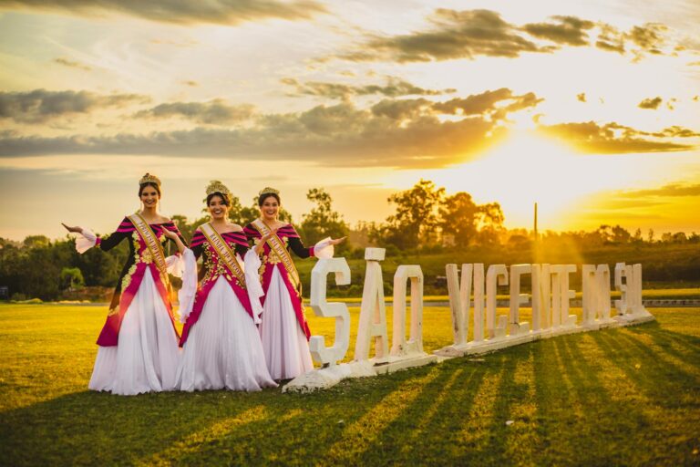 Elegant women in gowns posing by a São Vicente sign at sunset.