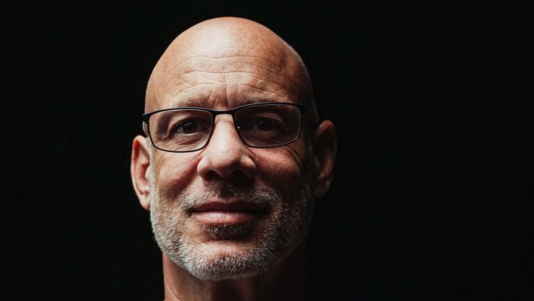 Close-up portrait of a bald man with glasses on a dark background.