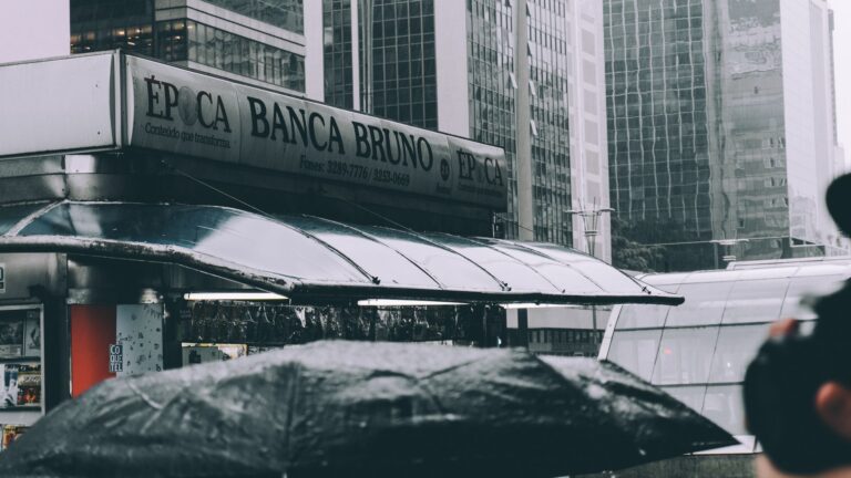 A street scene in a modern urban area featuring a magazine stand and high-rise buildings on a rainy day.