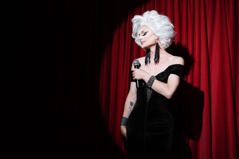Drag queen in a black dress with white wig singing on stage with a red curtain backdrop.