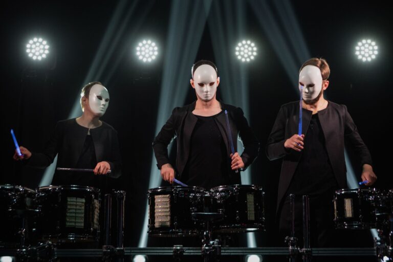A group of masked musicians playing drums under bright spotlights in a live concert setting.