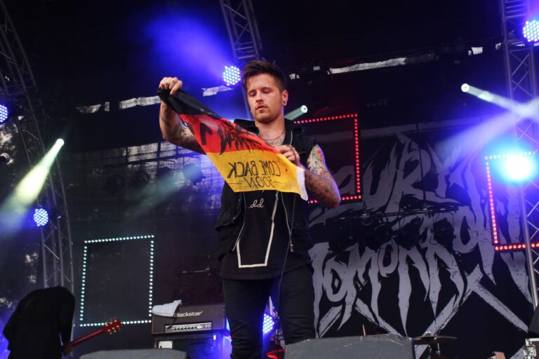 Tattooed musician on stage with guitar and flag at outdoor rock concert.
