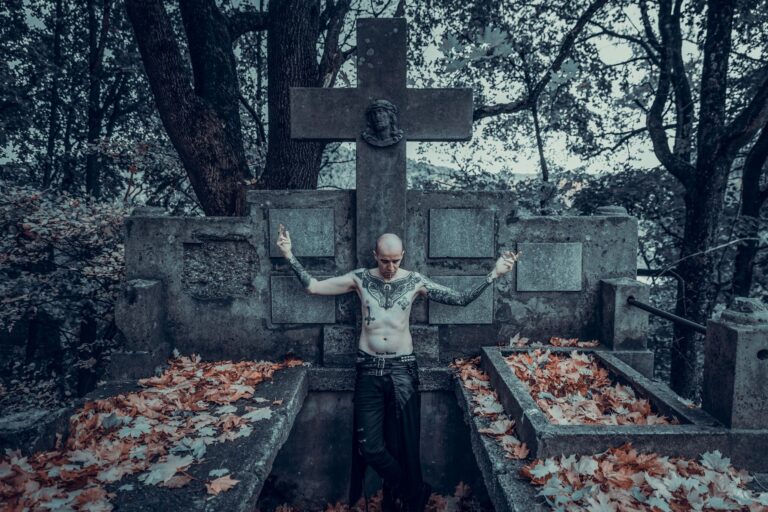 A shirtless, tattooed man standing in a cemetery, surrounded by autumn leaves and an ancient tombstone.