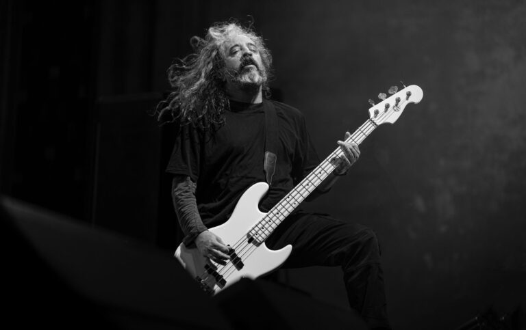Dynamic black and white photo of an adult male musician with long hair playing a bass guitar passionately.