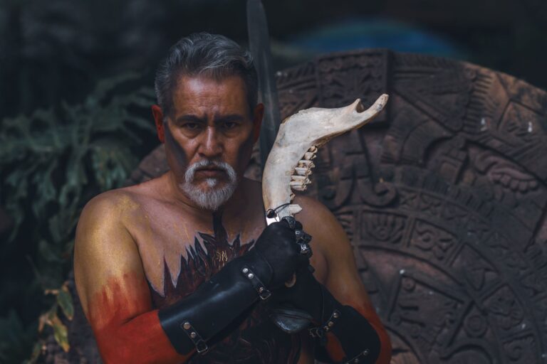 Portrait of tribal warrior with artistic body paint and animal horn against ancient symbol backdrop.