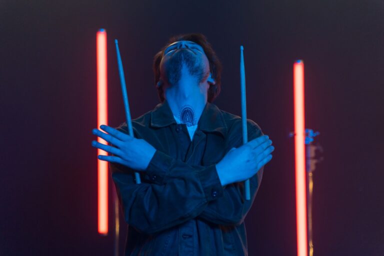 A man dramatically poses with drumsticks crossed under neon lights, conveying artistic expression.