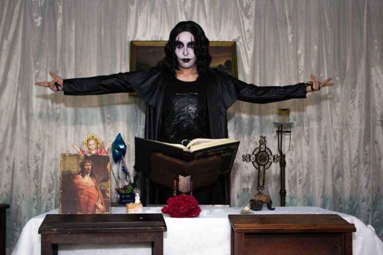 Person in gothic costume performing a dramatic scene indoors with religious symbols.