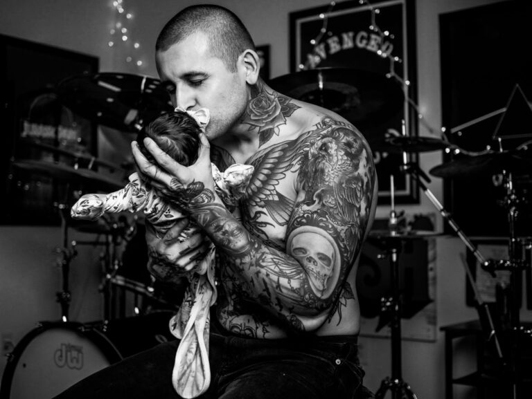 A tattooed man lovingly cradles a newborn baby in a music studio, exuding warmth and care.