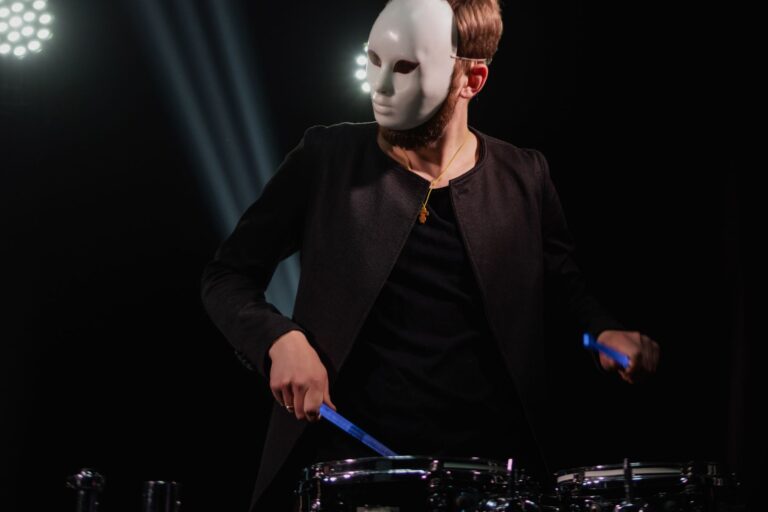 A drummer wearing a white mask plays energetically on stage under spotlights, creating a mysterious atmosphere.